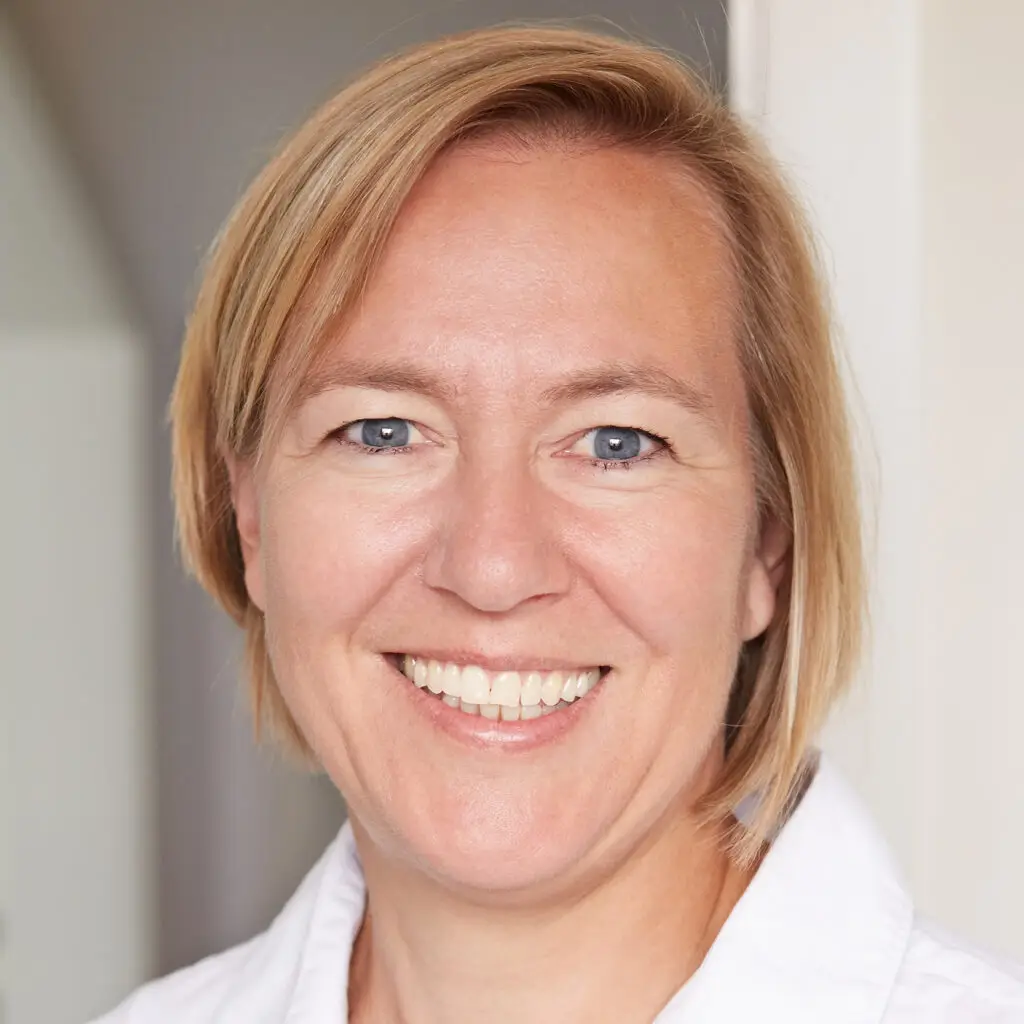 A person with short blonde hair from the Travel Services Team is smiling at the camera. They are wearing a white shirt, standing against a plain interior wall.