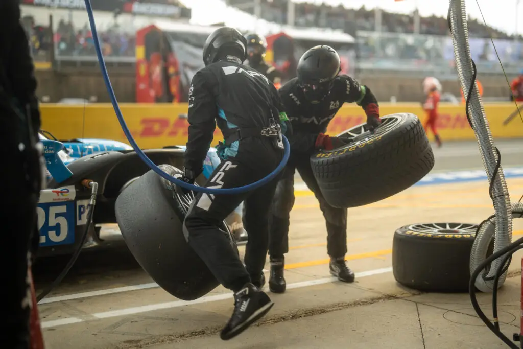 Amid the buzzing motorsport atmosphere, two pit crew members in black uniforms and helmets expertly change tires on a race car. As one member removes a tire, the other deftly prepares to install a new one, showcasing seamless cooperation in this high-speed event.