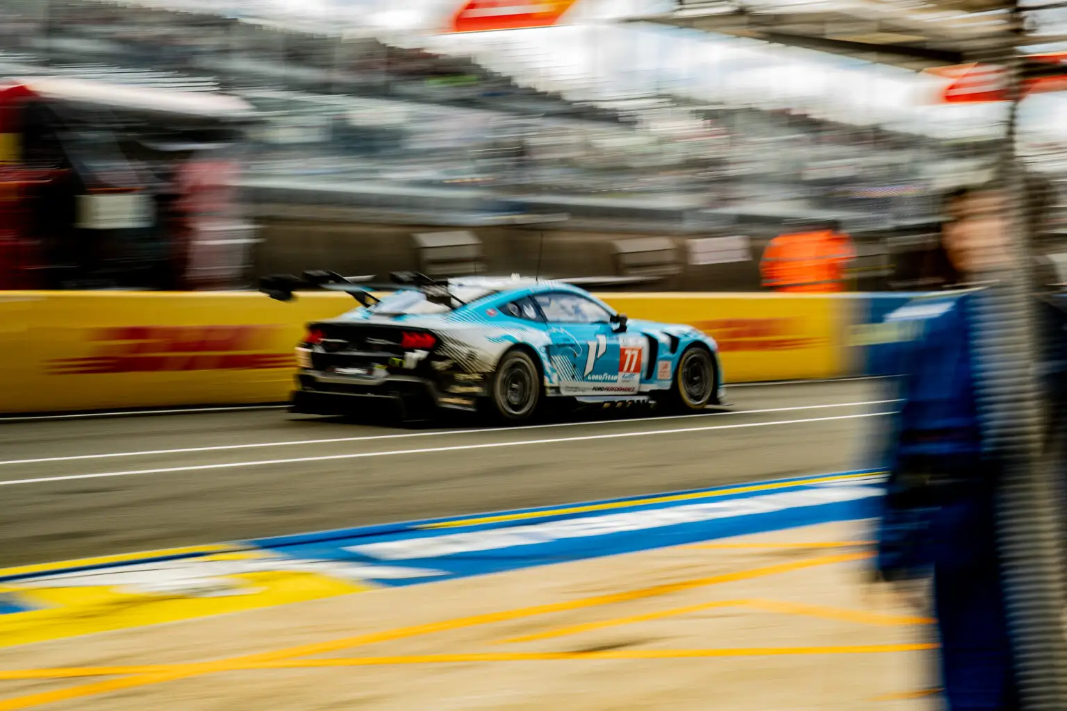 A silver race car, marked with number 11, streaks down the pit lane amidst the adrenaline-fueled energy of a motorsport event. Yellow safety barriers and indistinct figures blur into the background, epitomizing a seamlessly orchestrated race experience.