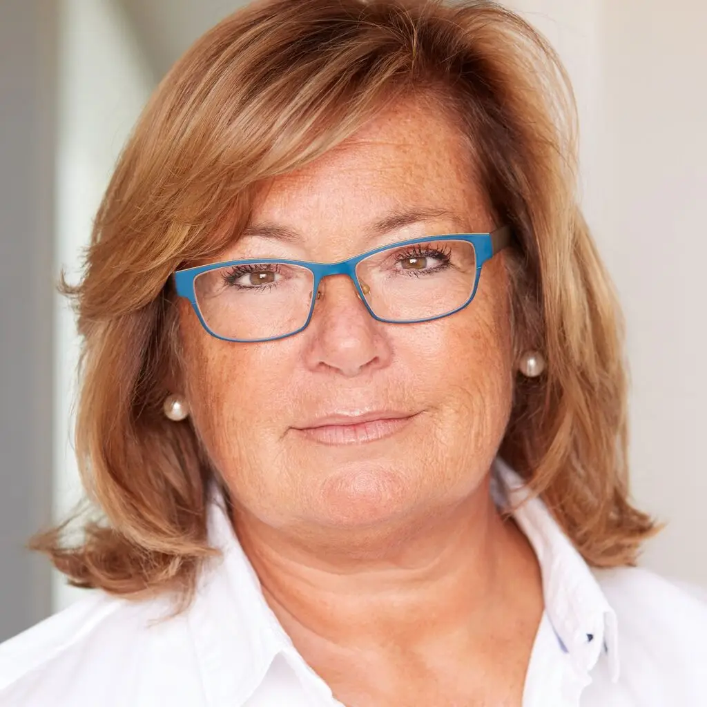 A middle-aged woman with shoulder-length blond hair and wearing blue glasses looks at the camera, embodying the professionalism of the Travel Services Team. Dressed in a white collared shirt and pearl earrings, she stands against a softly blurred background.