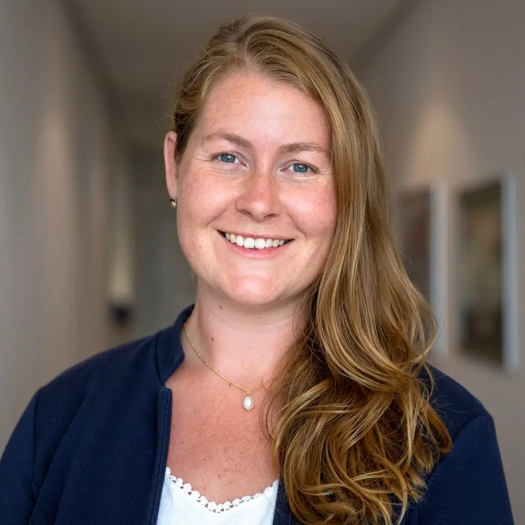 A woman with long, wavy hair smiles in a hallway, embodying the warm welcome typical of our Travel Services Team. She wears a dark jacket over a white top and a single pendant necklace. Soft lighting and blurred wall art create a calm atmosphere.