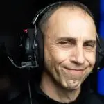 A man wearing headphones with a microphone smiles slightly. In the dark environment, a faint blue light suggests he's coordinating travel logistics. His expression appears calm and content, as if navigating the world from his seat.