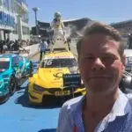 A man takes a selfie in front of parked race cars, capturing the thrilling atmosphere of motorsport. A person in racing gear celebrates on top of a yellow car with the number 16. In the background, a crowd gathers by a building, adding to the travel adventure vibe.