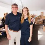 A man and a woman are standing side by side, smiling, inside a large tent with boxes stacked on tables in the background. Both are wearing dark polo shirts and light pants, looking like professionals managing travel logistics for a team event.